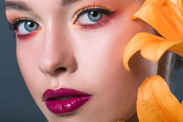 Retrato de cerca de la hermosa mujer joven con maquillaje de moda y flores de lirio naranja mirando a la cámara en gris - foto de stock