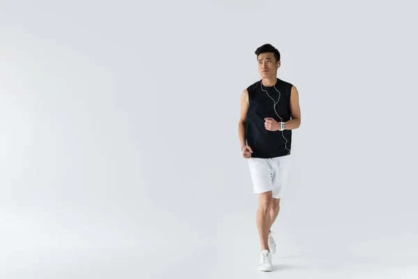 Ernsthafter junger asiatischer Sportler mit Kopfhörern, die auf grauem Hintergrund laufen — Stockfoto