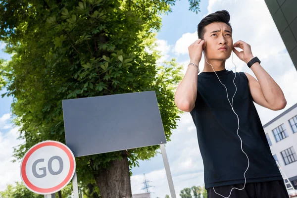 Низкий угол зрения серьезный азиатский спортсмен надевает наушники на улице с пустыми вывесками и дорожным знаком с надписью идти — стоковое фото
