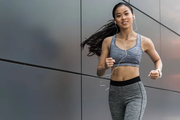 Усміхнена молода азіатська спортсменка в навушниках, що працюють на міській вулиці — стокове фото