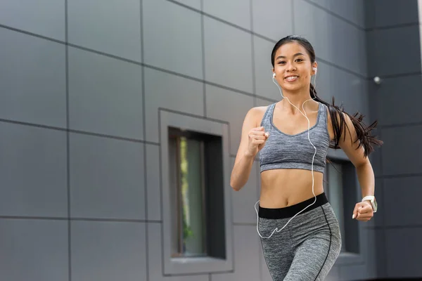 Smiling young asian sportswoman in earphones running at city street — Stock Photo