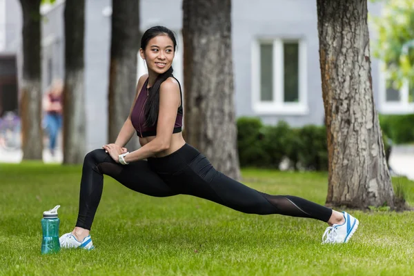 Sorrindo asiático feminino atleta alongamento perto esporte garrafa de água na grama no parque — Fotografia de Stock