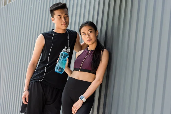 Sonriente joven asiático deportista con auriculares sobre el cuello de pie cerca de sportswoman con deporte botella de agua en la calle - foto de stock