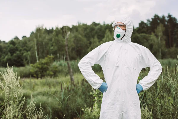 Fiducioso scienziato maschio in maschera protettiva, googles e tuta in piedi con le mani sulla vita nel prato — Foto stock