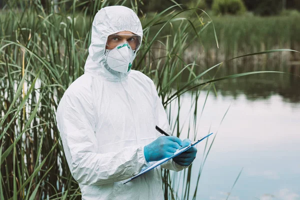 Male scientist in protective mask and suit writing in clipboard near water outdoors — Stock Photo