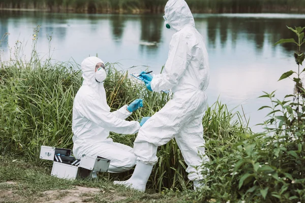 Científica en máscara protectora y traje escrito en portapapeles mientras su colega pone muestra de agua - foto de stock