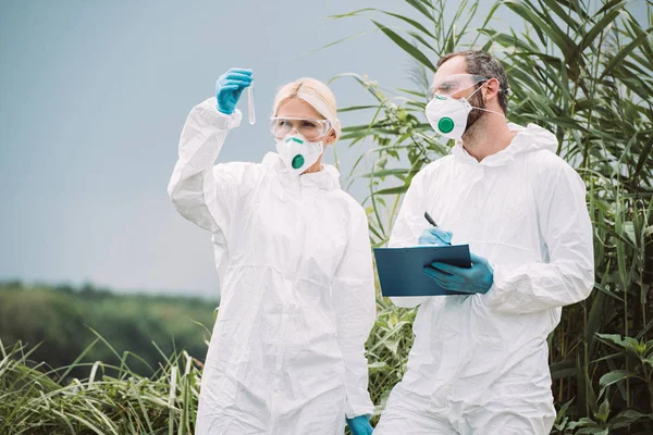 Mâle scientifique en combinaison de protection et masque écrit dans le presse-papiers tandis que son collègue femme examinant échantillon d'eau en fiole d'essai à l'extérieur — Photo de stock