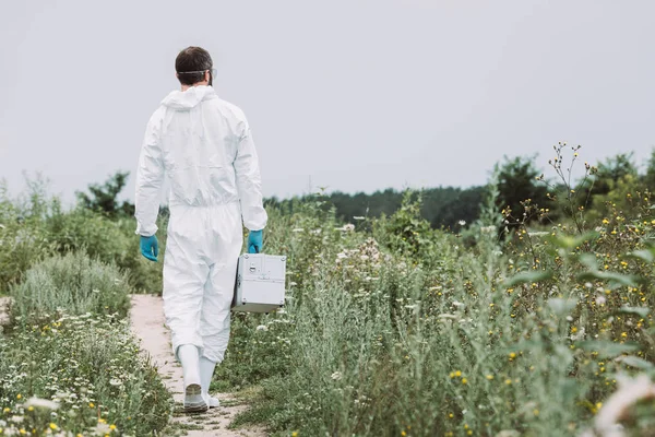 Rear view of male scientist in protective suit walking with working suitcase in meadow — Stock Photo
