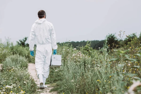 Vue arrière du scientifique masculin en combinaison de protection marchant avec valise de travail dans la prairie — Photo de stock