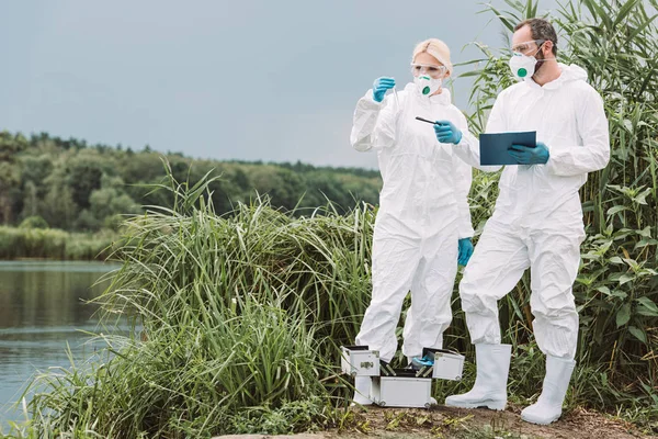 Male scientist in protective suit and mask writing in clipboard and pointing while his female colleague examining sample of water in test flask outdoors — Stock Photo