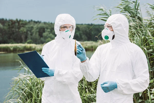 Male scientist in protective mask and suit holding fish by tweezers while his female colleague writing in clipboard outdoors — Stock Photo