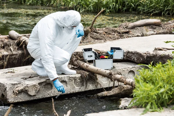 Científico masculino en traje protector y guantes de látex tomando muestra de agua en frasco de prueba al aire libre — Stock Photo