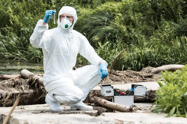 Male scientist in protective mask and suit examining sample of water in test flask outdoors — Stock Photo