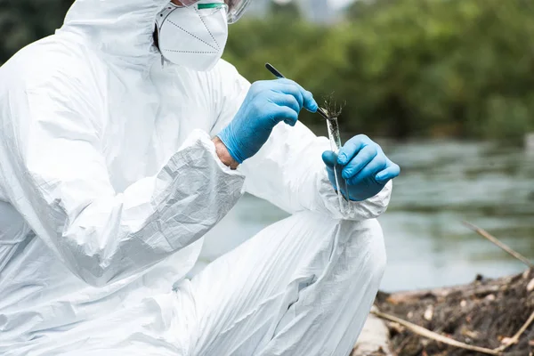 Imagen recortada de científico masculino en traje protector y máscara poniendo muestra de tierra por pinzas en frasco de prueba al aire libre — Stock Photo