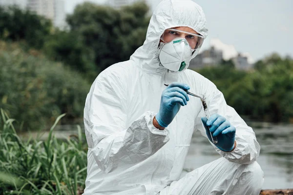 Científico masculino serio en traje protector y máscara poniendo muestra de suelo por pinzas en frasco de prueba al aire libre - foto de stock