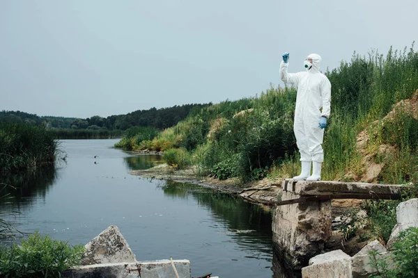 Male scientist in protective mask and suit looking at sample of water in test flask outdoors — Stock Photo