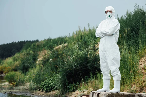Male scientist in protective suit and mask standing near water outdoors — Stock Photo