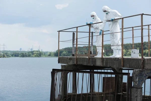 Male scientist in protective suit and mask pointing at water to female colleague with laptop outdoors — Stock Photo