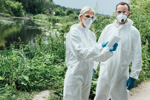 Female and male scientists in protective masks and suits working with digital tablet and looking at camera outdoors — Stock Photo