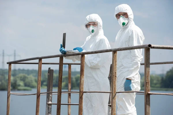 Two scientists in protective suits and masks working with laptop and looking at camera near river outdoors — Stock Photo