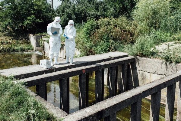 Two scientists in protective suits and masks working with laptop near sewerage — Stock Photo