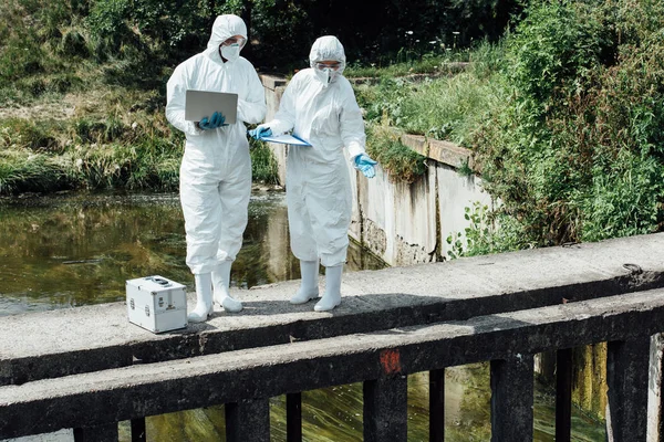 Female scientist pointing on water of sewerage to male colleague with laptop — Stock Photo