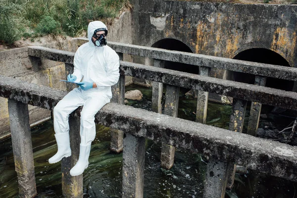 Male scientist in protective mask and suit writing in clipboard while sitting near sewerage — Stock Photo