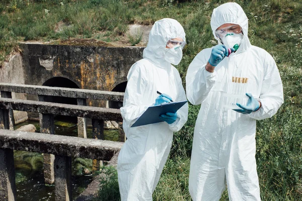 Científico masculino poniendo la muestra de agua en frasco de prueba mientras su colega escribiendo en portapapeles cerca de alcantarillado - foto de stock