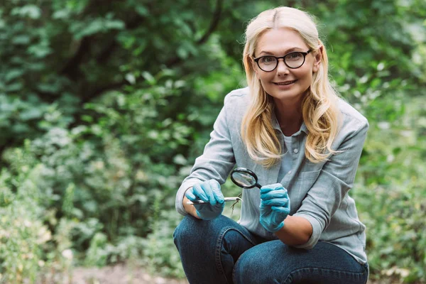 Femme scientifique en gants de latex assise avec brindille dans une pince à épiler et loupe dans la forêt — Photo de stock