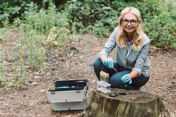 Smiling female scientist putting twig by tweezers in test flask in forest — Stock Photo