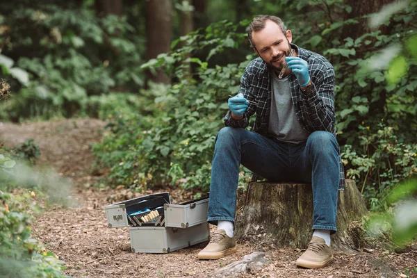 Científico masculino sonriente en guantes de látex tomando muestra de hoja seca en frasco de prueba en el bosque - foto de stock
