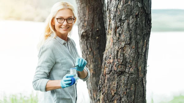 Smiling female scientist in eyeglasses putting sample by tweezers in jar near trees outdoors — Stock Photo