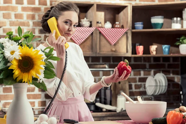 Lächelnde erwachsene Hausfrau, die mit einem kabelgebundenen Retro-Telefon spricht und in die Küche in die Kamera schaut — Stockfoto