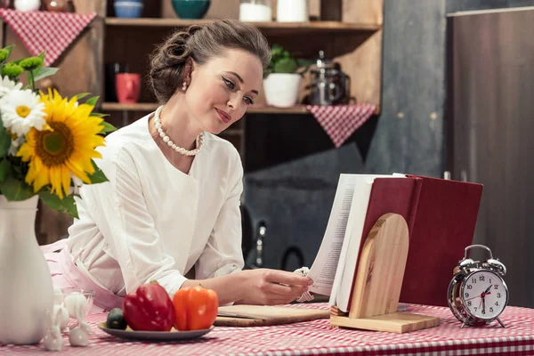 Sonriente atractivo ama de casa en la ropa vintage lectura receta libro en la cocina - foto de stock