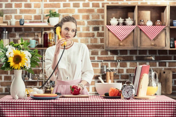 Красивая взрослая домохозяйка разговаривает по телефону во время приготовления еды на кухне — стоковое фото