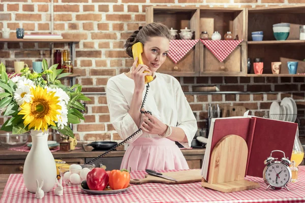 Hermosa ama de casa adulta hablando por teléfono y mirando a la cámara durante la preparación de la comida en la cocina - foto de stock