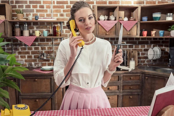 Sérieux adulte ménagère avec couteau parler par téléphone filaire jaune à la cuisine — Photo de stock