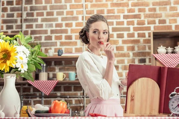 Красивая взрослая домохозяйка ест сырой перец и смотрит на кухню — стоковое фото
