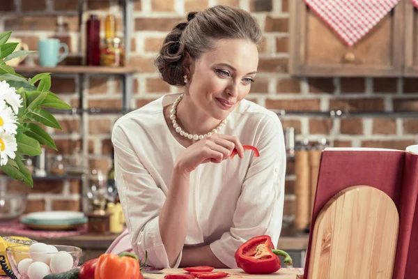 Lächelnde erwachsene Hausfrau liest Rezeptbuch und probiert frisches Gemüse in der Küche — Stockfoto