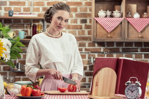 Приваблива доросла домогосподарка в старовинному одязі, що ріже помідор і дивиться на камеру на кухні — стокове фото