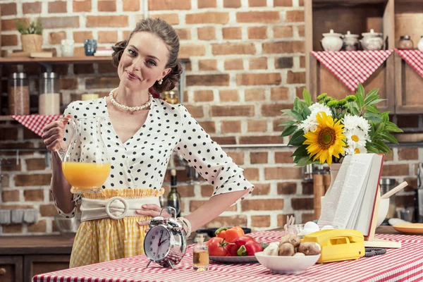 Happy adult housewife holding jug of orange juice and looking at camera at kitchen — Stock Photo