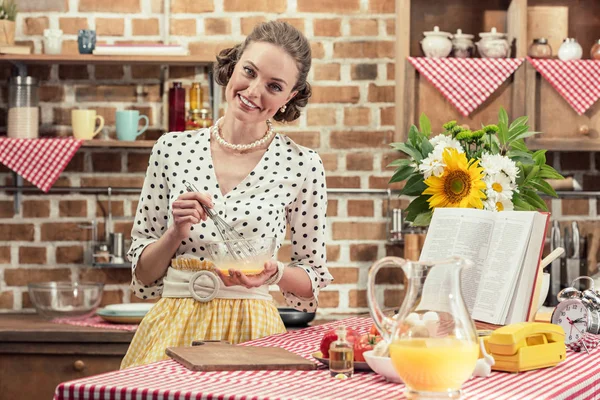 Glückliche erwachsene Hausfrau, die Eier für Omelette schlägt und in die Küche in die Kamera blickt — Stockfoto