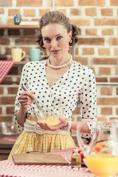 Attrayant adulte femme au foyer fouettant des œufs pour omelette et regardant la caméra à la cuisine — Photo de stock