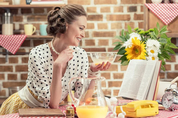 Sorridente casalinga adulta con ciotola lettura libro di ricette in cucina — Foto stock