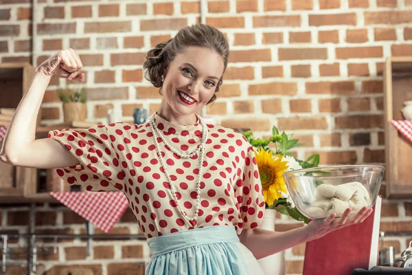 Casalinga adulta sorridente che tiene la boccia con pasta e mostra muscoli a macchina fotografica a cucina — Foto stock