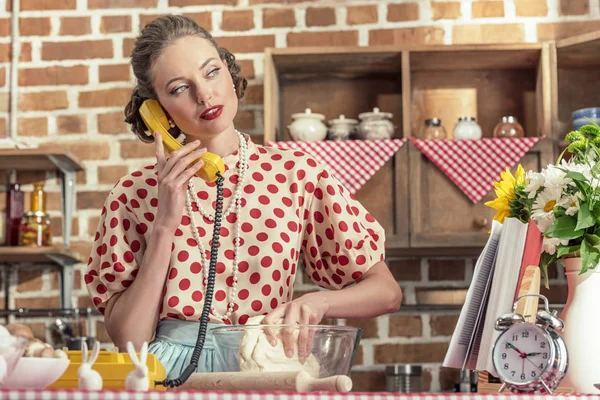 Atractiva ama de casa adulta hablando por teléfono y amasando masa en la cocina - foto de stock