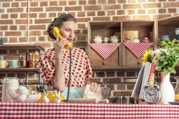 Beautiful adult housewife talking by phone and kneading dough at kitchen — Stock Photo