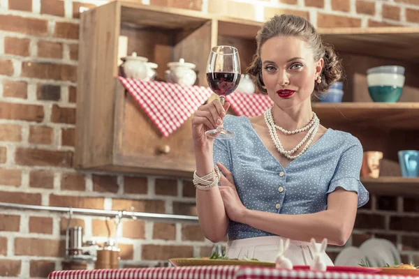 Красивая взрослая домохозяйка с бокалом красного вина, смотрящая в камеру на кухне — стоковое фото