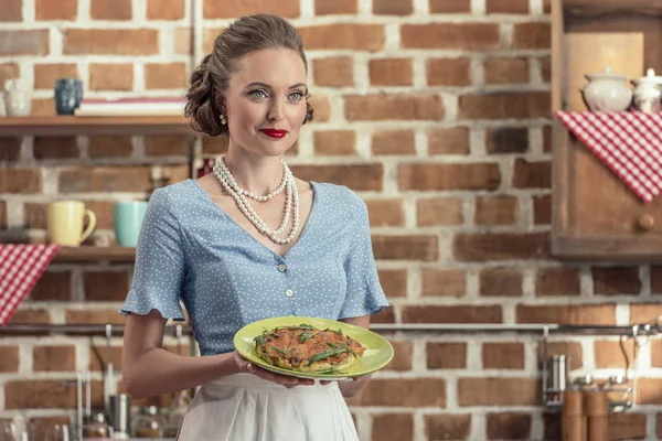 Sonriente ama de casa adulta en ropa vintage sosteniendo plato con pastel de setas en la cocina - foto de stock