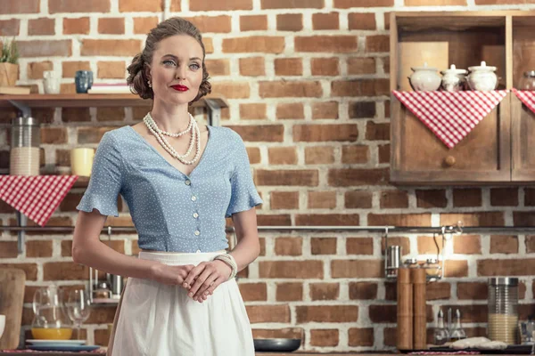 Улыбающаяся взрослая домохозяйка в винтажной одежде стоит на кухне и смотрит в сторону — стоковое фото
